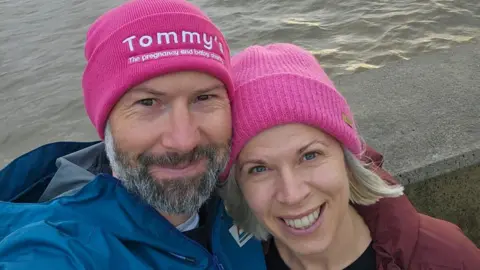 A bearded man wearing a bright pink beanie hat with Tommy's the pregnancy and baby charity written on the front takes a selfie with a blonde woman also wearing a bright pink hat. They are stood in front of the sea