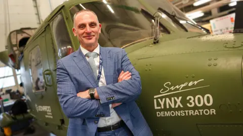 Yeovil College A man in a blue suit and white shirt is looking at the camera with his arms folded, smiling. He is stood infront of a green helicopter, bearing the name 'lynx 300'.