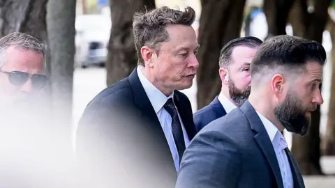 Elon Musk, in a dark suit and tie with a crisp white shirt and pursed lips, arrives at federal court on March 4, 2026 in San Francisco, California. 