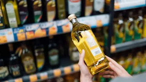 Getty Images Someone inspects a bottle of olive oil in front of supermarket shelves containing multiple brands of olive oil.