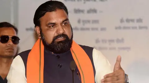 Samrat Choudhary - wearing a sleeveless vest over a cream-coloured kurta and sporting a saffron stole - speaks at a workshop at Hotel Taj City Centre in Bihar's capital city Patna on 4 April 2026. Behind him there is a bodyguard wearing sunglasses.