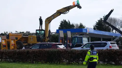 An Irish police officer is looking toward a protester standing on a crane beside a petrol station. An Irish flag is being flown on top of the crane.