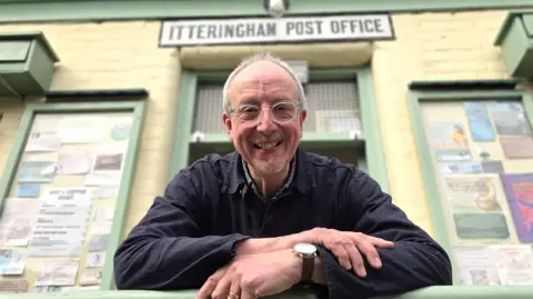 Mike Hemsley is leaning in front of a shop. He's wearing a dark blue shirt and transparently-rimmed glasses. Behind him is the 'Itteringham Post Office' sign.