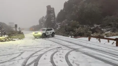 An emergency services car parked across a snow-covered rural road in fog