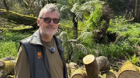 A man in National Trust branded gardening clothes smiles at the camera, behind him is a tree that was downed by Storm Goretti and parts of another tree cut up and ready to be taken away 