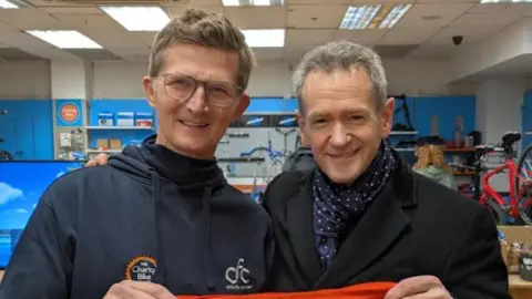 Alexander Armstrong is on the right and has grey hair with a black coat and blue and white scarf. He is holding one side of a red top that reads "one big thank you" in white writing. On the left is Michael who has brown hair and glasses and is wearing a blue navy hoodie, and is holding the other side of the top.