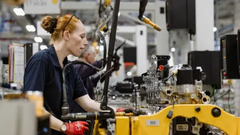 PA Media A woman with her hair tied in a bun wears a blue tshirt while using tools on an engine on a factory floor.