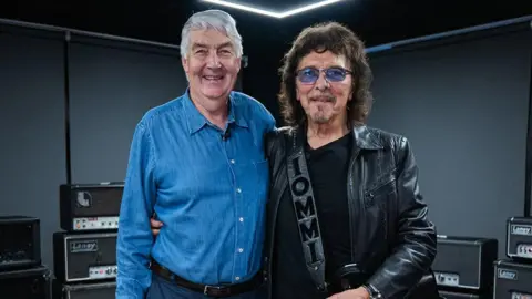 Lyndon Laney, with short grey hair, is wearing a blue denim shirt. He has his arm around Tony Iommi who is wearing dark glasses, a leather jacket and a guitar strap with Iommi printed on it.