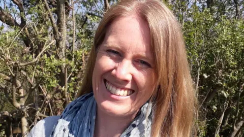A woman with blonde hair, wearing a white top and a scarf, smiles at the camera. There are trees and shrubbery behind her.