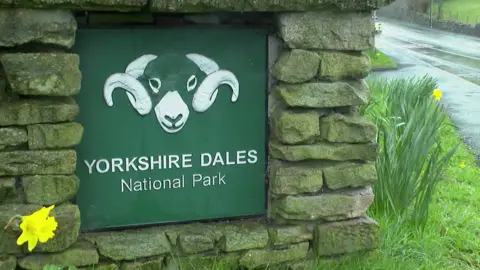 The image shows a roadside sign marking the entrance to the Yorkshire Dales National Park. The sign is mounted within a low structure made of irregularly shaped stone blocks.