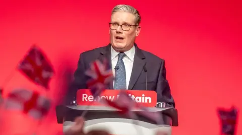 Starmer speaks at a podium in front of a red wall, with people waving Union Jacks in the crowd