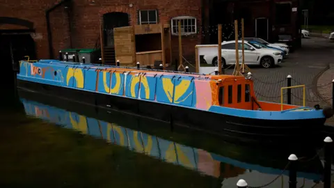 A canal boat which is painted blue with yellow circles, pink pint strokes and has a red and orange hull