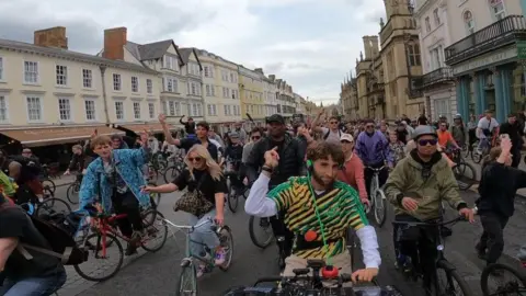 DJ Dom Whiting playing music while on his bike in Oxford. He is on the High Street. Hundreds of ravers are behind him on their bikes, following him. 