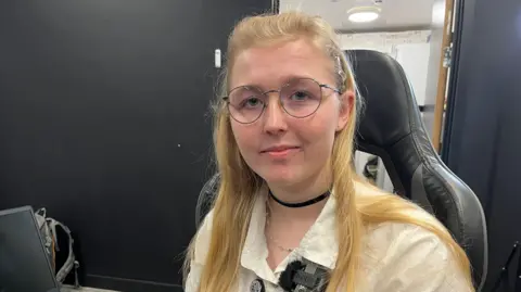 Ant Saddington/BBC A young woman with glasses and blonde hair who is looking at the camera. She is sitting down and is wearing a white shirt.
