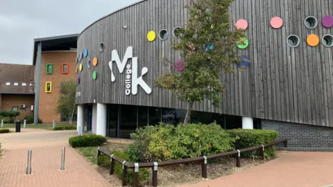 Sam Read/BBC The front of Milton Keynes College, which has a small tree and bushes outside as well as a pedestrianised area. The wooden-clad building has small round windows of different colours.