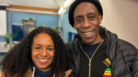 A picture of married couple Amanda Sterling-Bynoe and Derek Sterling smiling towards camera, at the family history masterclass. She has long black curly hair and he has a black hat on