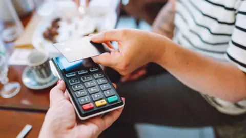 A woman wearing a white and blue striped top taps her bank card on a card reader to pay for food and drinks in a cafe. 