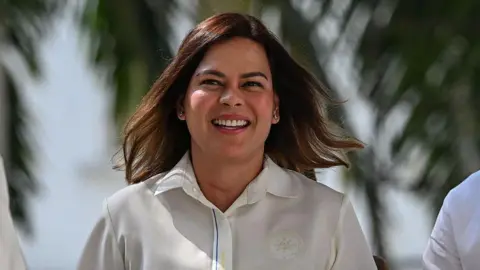 Getty Images Sara Duterte smiling in a white button-up shirt while standing in front of palm trees