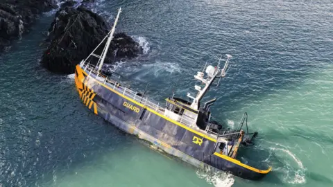 Wide drone shot shows the large navy and orange vessel sunk in the sea leaning on one side. On the left, black rocks and cliffs can be seen sitting right next to the front of the ship.