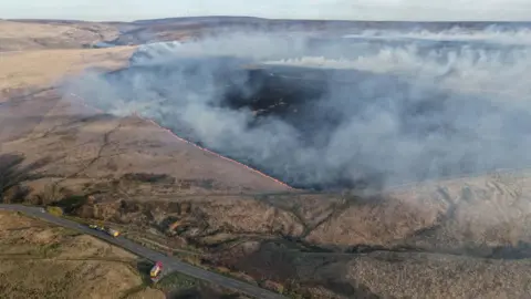 An aerial view showing a large expanse of moorland with smoke rising from the ground and orange flames around a section of blackened land