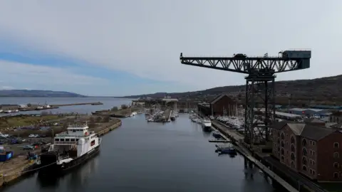 Aerial view of Greenock waterfront, showing a dock basin with boats, a large historic crane beside red‑brick warehouse buildings, and hills rising behind the River Clyde.
