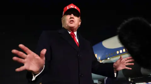Donald Trump pictured from below with his hands outstretched, wearing a red USA baseball cap, with a plane visible in the background