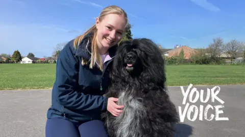 Megan Slack is kneeling on concrete holding a large furry black dog which has white hairs on its chest. Megan has long blonde hair and is wearing a blue tracksuit.