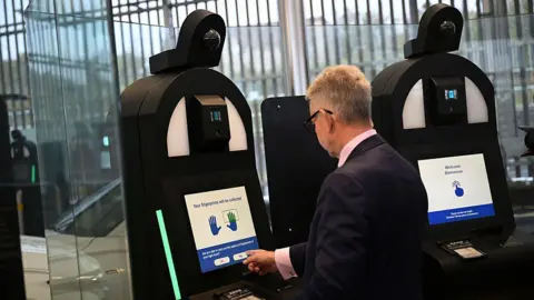 A man with grey hair and wearing a dark suit is entering his personal details into a biometric machine. 