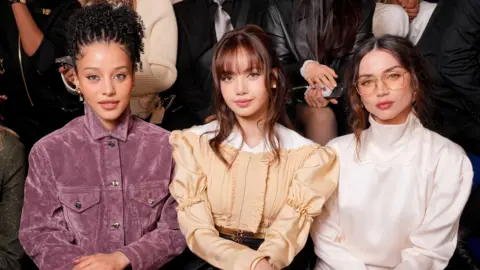Getty Images Chase Infiniti in a purple jacket, Lisa in a peach blouse, and Ana de Armas in a white high-necked top, at the Louis Vuitton fashion show