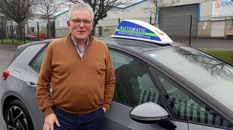 A man with white hair and glasses standing smiling in front of a learners car parked on a street by an industrial estate. He is wearing a brown top and navy bottoms.