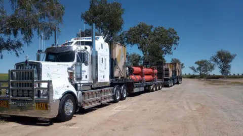 A road train on a dirt road with trees and blue skies in the background