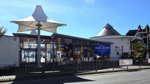 The North Norfolk Visitor Centre in Cromer. A relatively modern building, the main part of it is glass-fronted. There is a 'for lease' sign in front of it.