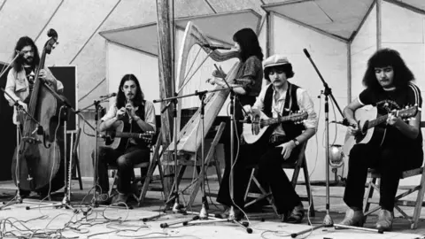 Getty Images Clannad performing in a studio playing instruments including a cello, a guitar, a harp and stringed instruments. Some of the band members are standing and some are seated. The image is in black and white.