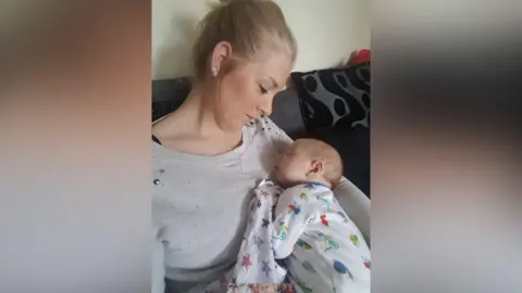 Laura sitting on a sofa and cuddling her tiny baby, who is in the crook of her arm asleep wearing a babygro with insects on it. Laura is looking down adoringly at him, wearing a grey jumper