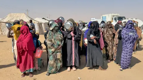 Ed Habershon / BBC A group of women in al-Dabbah wearing long flowing robes and holding receptacles stand outside in a desert-like setting. Behind them can be seen white tents and a lorry.