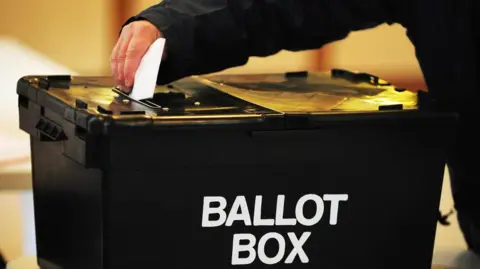 A voter puts his voting slip in a black ballot box. He is wearing a black coat.