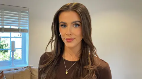 Charli Aitken - a woman - looks at the camera. She is standing in front of a sofa and a window. She has long brown hair and a brown top. She is wearing hoop earrings and a necklace with a pendant.