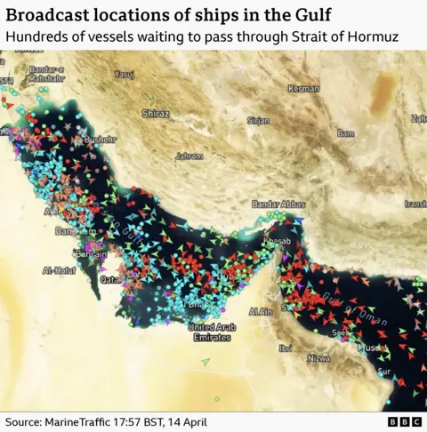 A satellite image of the Strait of Hormuz with ships marked with arrows. It shows hundreds of ships clustered by the strait waiting to pass. It is timestamped to 17:57 BST on 14 April