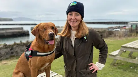 BBC A fox red Labrador is sitting on top of a picnic bench. One of his eyes has been sewn shut. His mouth is open and his tongue is sticking out. A woman has one hand on his back and the other hand on her hip. She's wearing a hat that says RSPB.
