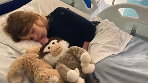 Winifred is pictured asleep in a hospital bed, holding two soft toys under a blanket, with hospital rails visible around the bed.
