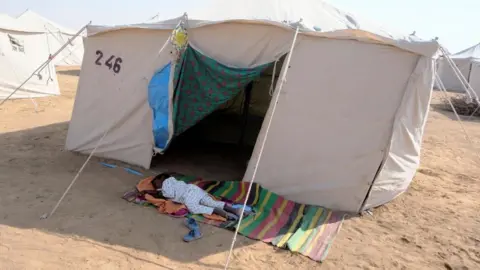 Reuters A child from el-Fasher sleeps on the ground on a rug outside a white tent near the displacement camp in al-Dabbah, Sudan, 12 November 2025.
