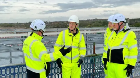 Four men wearing fluro yellow jackets and trousers and white safety helmets stand in front of a vast field of steel beams.