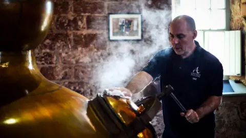 A man in a blue T-shirt stands next to a copper whisky still. There is steam coming out of a door on its side. 