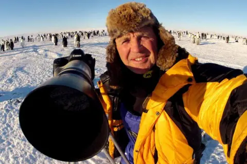 Doug Allan Doug Allan in a yellow jacket and furry hat with a large camera with hundreds of penguins in a frozen landscape in the distance behind him