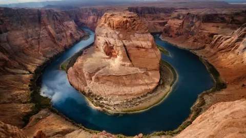 An aerial shot of a horse shoe bend of the Colorado River - the water is dark blue and the rock rusty brown.