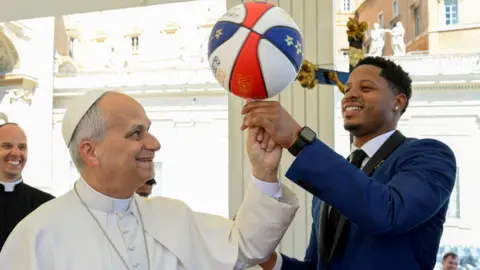 Pope spinning a basketball on his finger with the help of a Harlem Globetrotter player