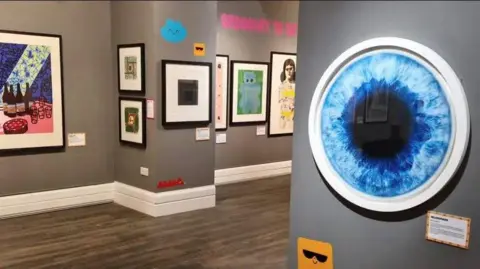 A gallery space with grey walls that has colourful artworks hanging on the walls.