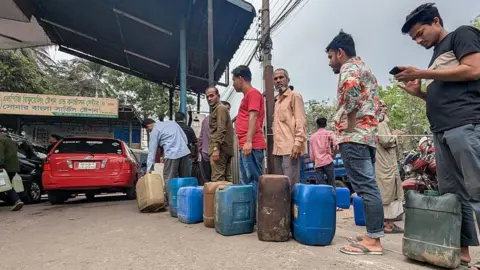 Motorists wait in line to purchase fuel at a petrol station in the Mohammadpur area of Dhaka, Bangladesh. Several people are standing in a queue with various petrol cans waiting to be filled