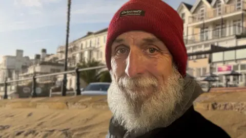 A shot of Sandown's promenade and its buildings from the beach. Ian Boyd is in front of the railings. He is wearing a red hat and a grey/black coat and has a long grey beard. 