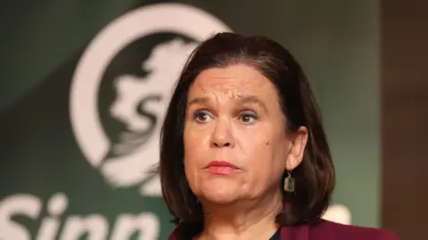 Mary Lou McDonald is looking off to the left. She has short, brown bobbed hair and you can just about see the top of her maroon jacket. She is wearing pink lipstick and earrings.